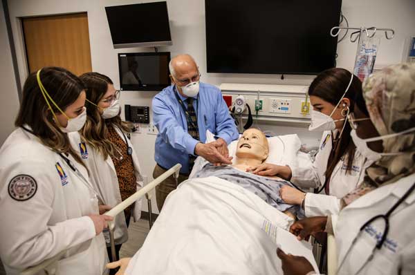 An instructor speaks to a group of students gathered around a simulated patient.