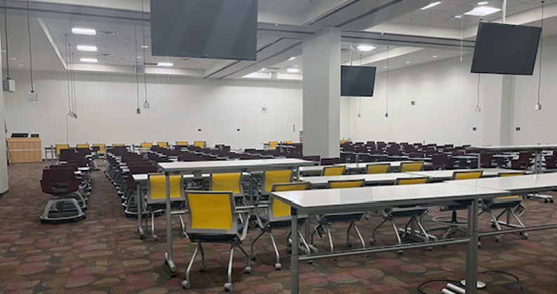 Large classroom with long tables, chairs and large flatscreen monitors and a front podium used for debriefing after Sim Center exercises