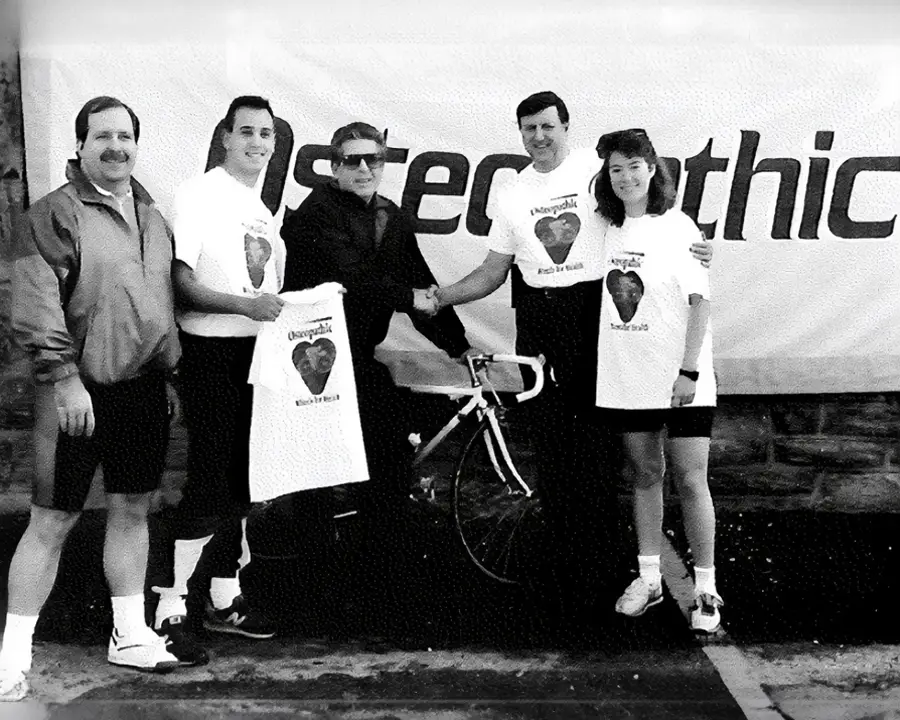 Black and white photo of Dean Finkelstein sitting on a bike and smiling with students during a PCOM community event in 1990