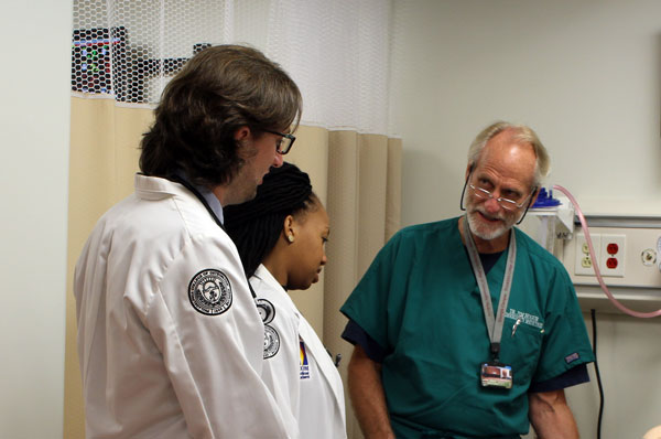 A doctor dressed in scrubs speaks with two medical students in an examination room.