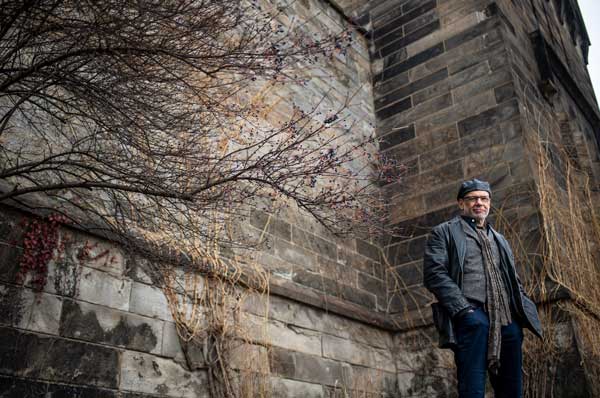 Dr. Gregory McDonald standing in front of a stone wall wearing a black leather jacket and a black cap.