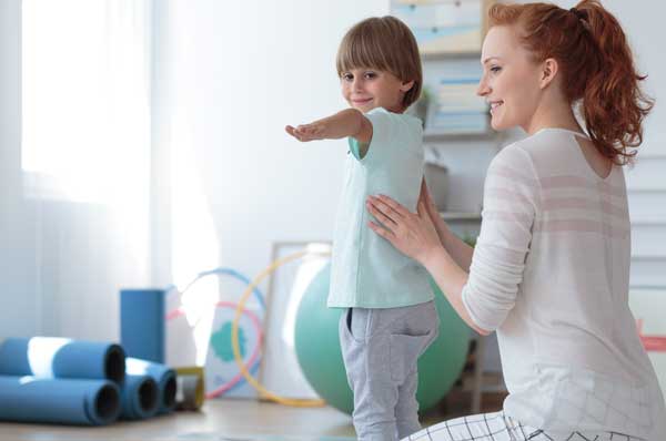 A pediatric physical therapist works with a child who is standing with his arms extended.