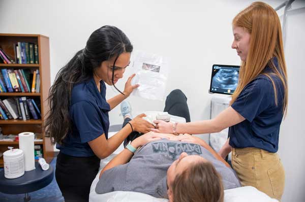 Two female students use an ultrasound device on a patient.