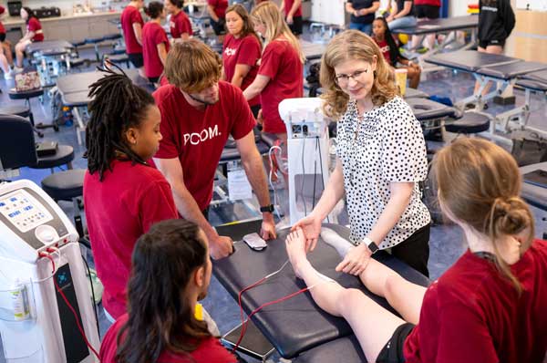 A female physical therapy instructor demonstrates a technique while surrounded by students in a classroom.