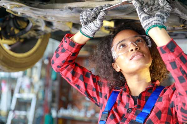A Black female mechanic works on the underside of a vehicle.