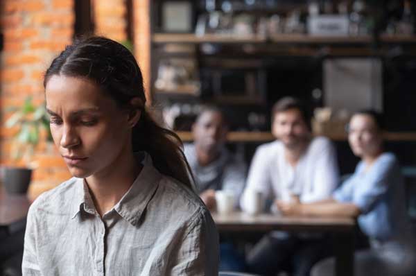 A sad woman frowns while people laugh in the background.