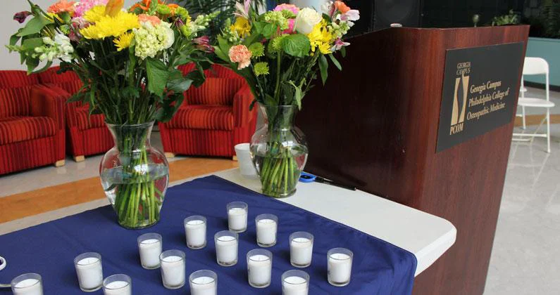 Two bouquets of colorful flowers in glass vases and several small, white candles sit on a table with a blue tablecloth. 