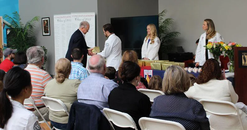 Seated guests watch as a PCOM Georgia student in a white coat hands a box of cremains to a man in a black suit. 