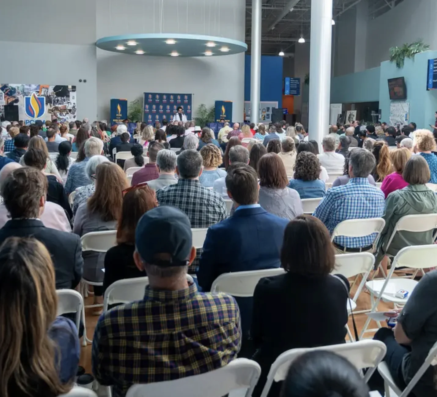 A large audience seated in a hall watch as a speaker stands at a podium