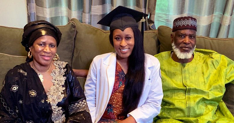PCOM physician assistant program graduate Bintou Berete, MS/PA '20, wears her cap and white coat and smiles on a couch with her parents