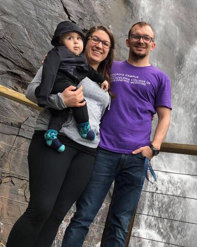 Nathan Ambrose, MS/PA '20, wears his PCOM t-shirt and poses with his wife and child in front of a waterfall.