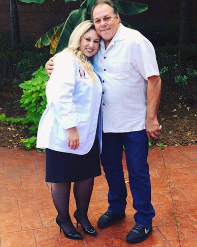 Samantha Sabada wears her pharmacist white coat and poses with her father in front of PCOM Georgia