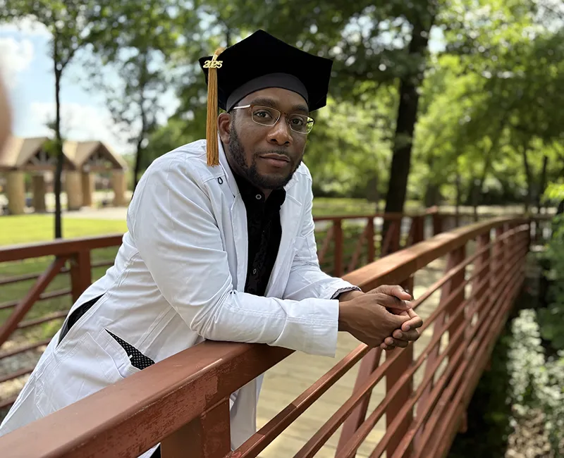 PCOM DO grad Corey Nick Brisseau, DO '25, looks thoughtful as he stands on a small bridge in a park wearing a white suit and his graduate cap