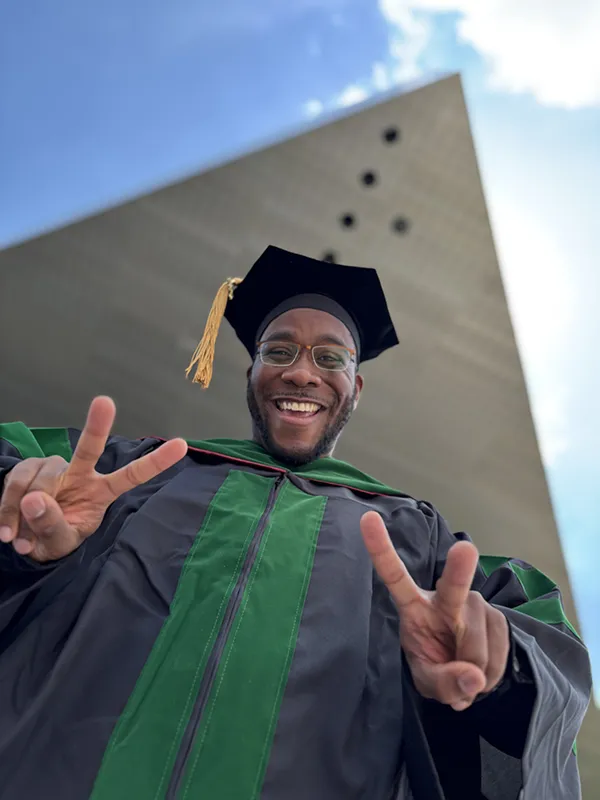 PCOM med school graduate Corey Nick Brisseau, DO '25, smiles and gives two peace signs in front of an architectural structrure