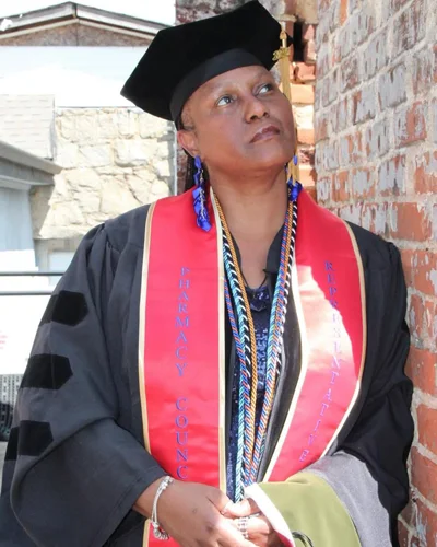 Makeda Lovelace, PharmD ’25, leans against brick wall while wearing commencement regalia