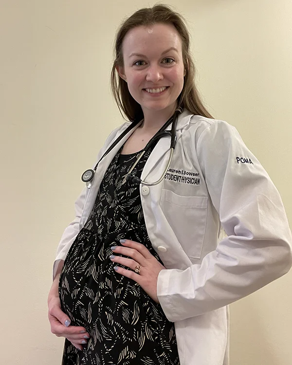 PCOM graduate Lauren Bowser, DO '26, smiling in her student physician white coat while holding her baby bump