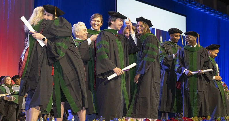 PCOM graduates wearing caps, gowns and regalia hugging and celebrating on the stage during a commencement ceremony