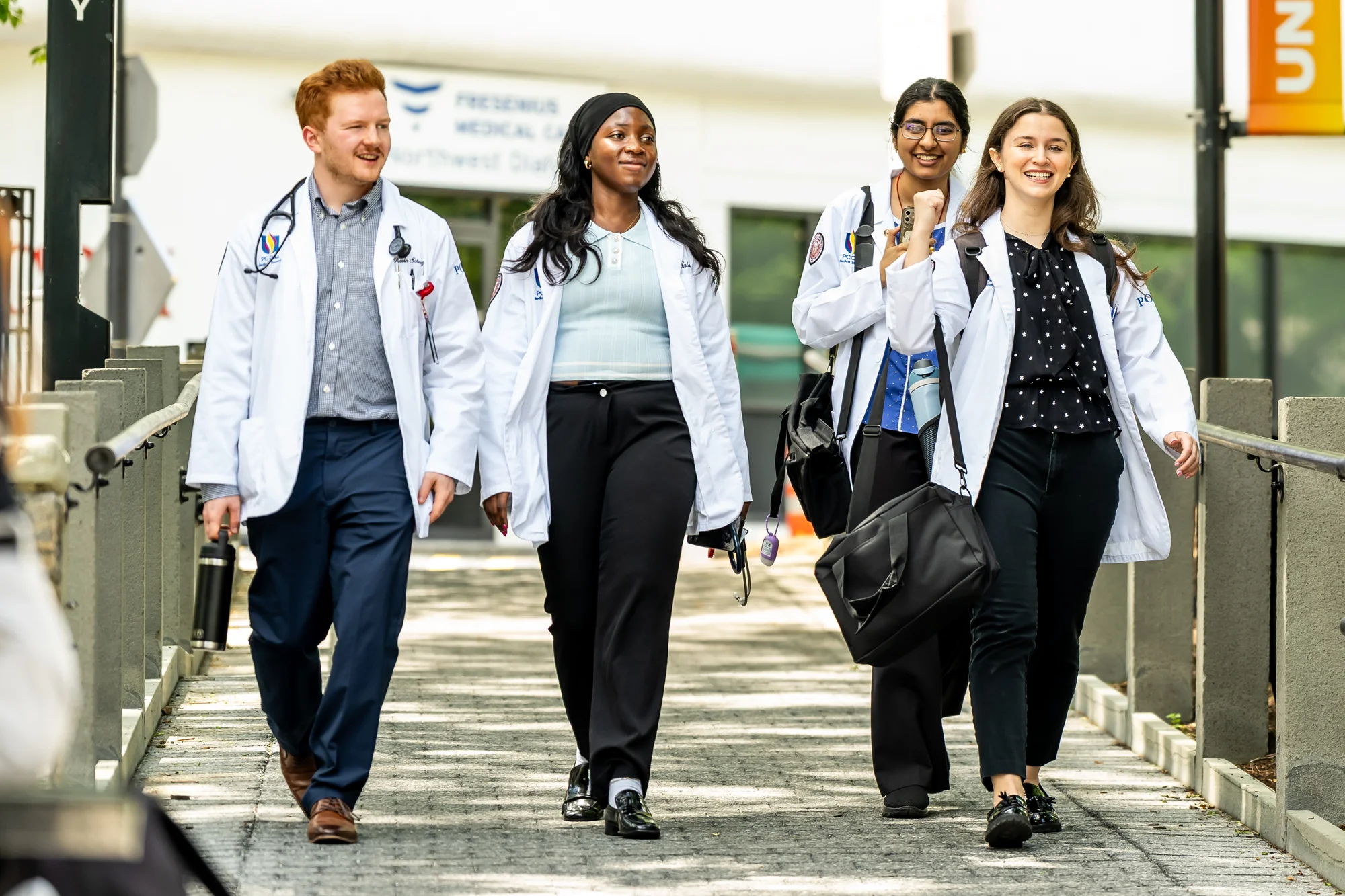 Medical students in white coats walk and talk on a pathway outside Philadelphia College of Osteopathic Medicine's main campus