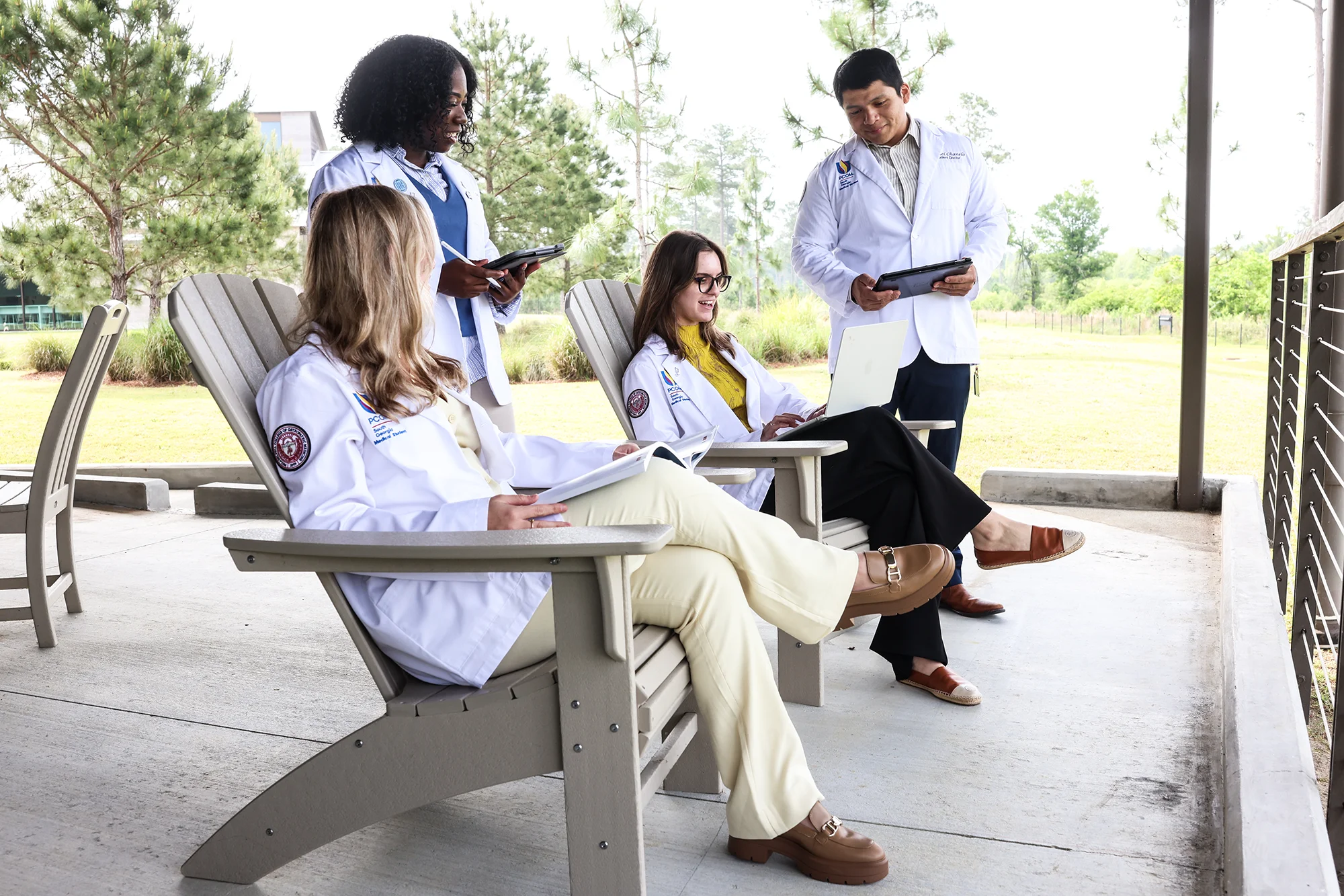 Medical students in white coats study outside with tablets and books on the rear porch of PCOM South Georgia