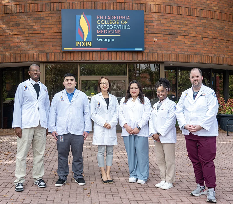 PCOM Georgia student researchers and faculty stand in front of PCOM Georgia's main campus front doors