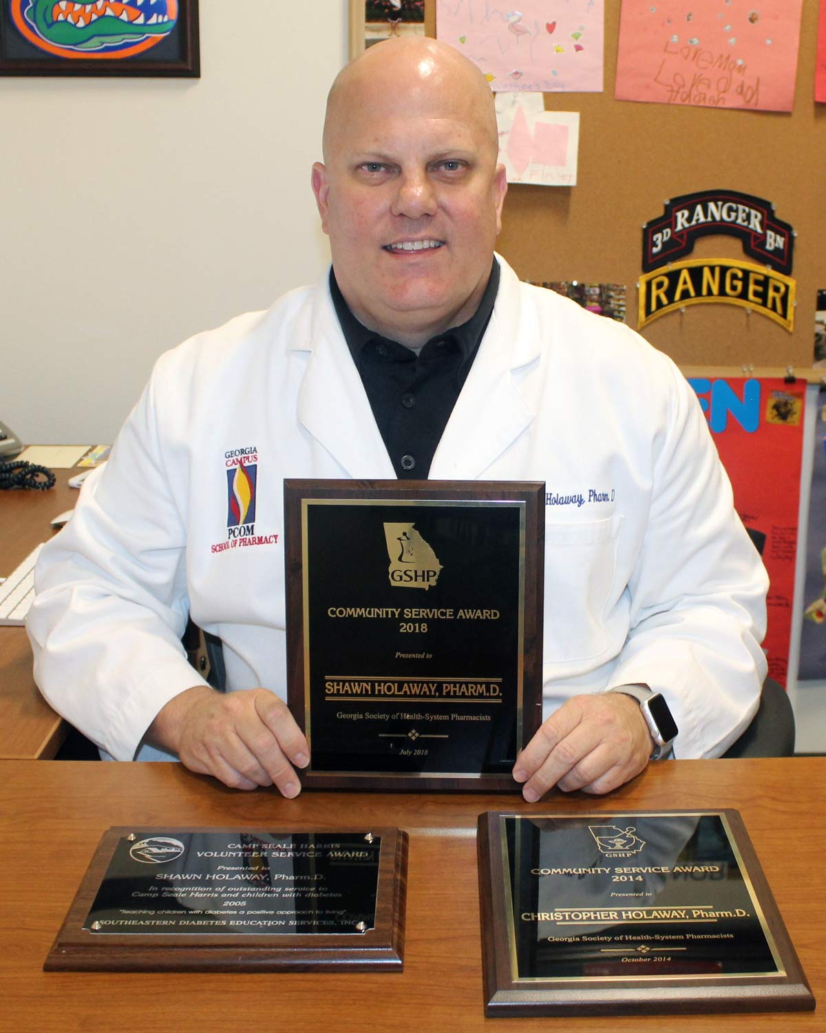 Photograph of Christopher Holaway, PharmD, at his office desk holding his community service award.