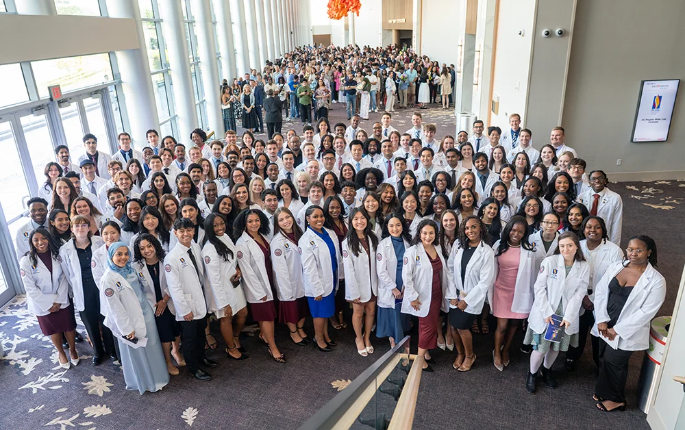 A wide aerial group shot of dozens of PCOM PA Studies students smiling in their new white coats