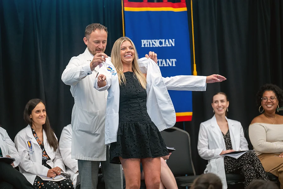 A female physician assistant student smiles as a faculty member helps her put on the student physician white coat