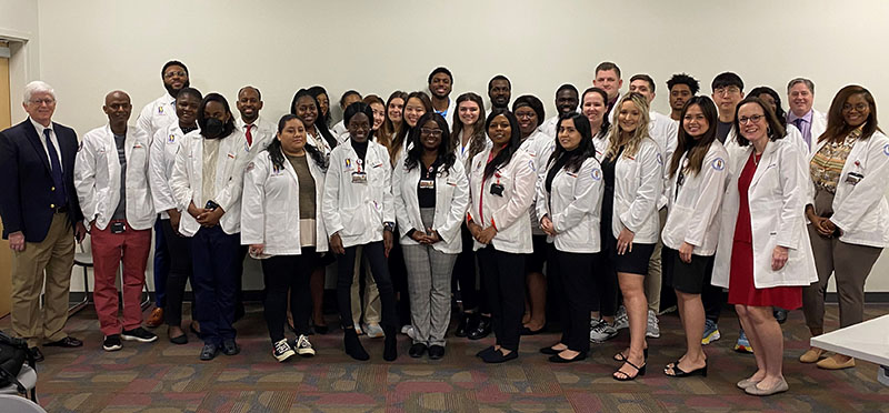 PCOM Georgia pharmacy students pose in their white coats with faculty in a classroom during the 2023 virtual Day at the Dome event.
