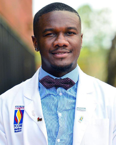 Professional headshot photo of Aldwin Soumare (DO '22) wearing his physician white coat in front of the PCOM Georgia entrance sign