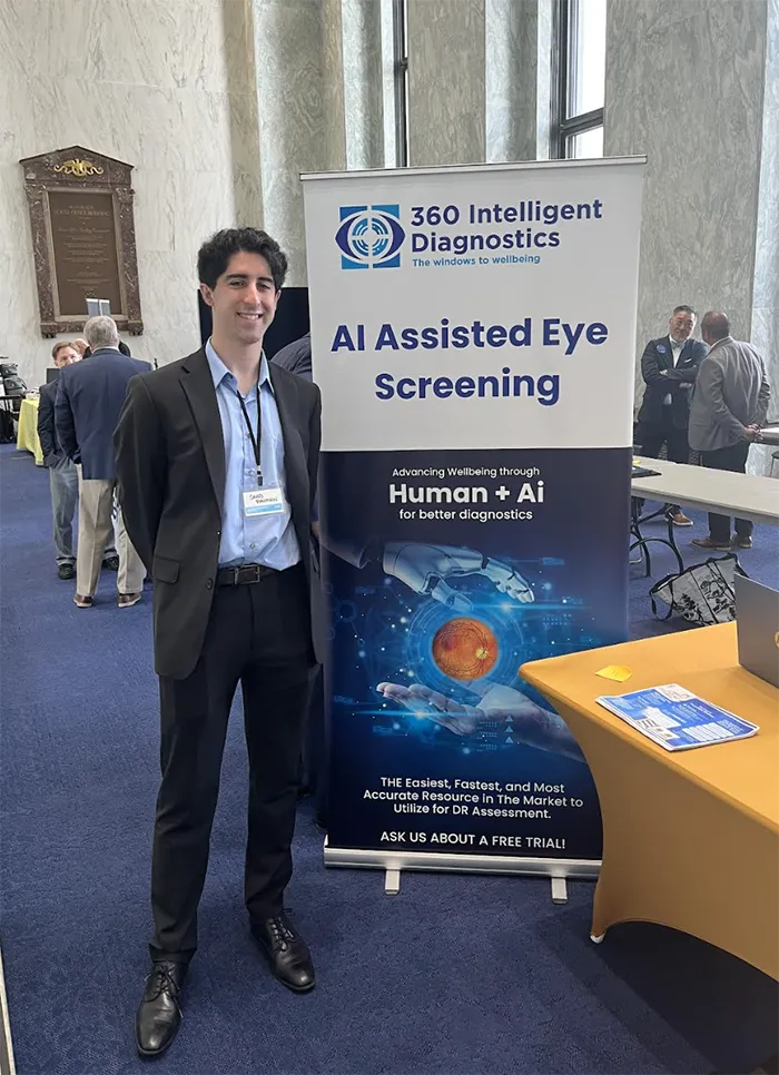 PCOM medical student Saad Rahman smiles in front of his company's sign offering AI-assisted eye disease diagnostics at a health technology conference at the US Capitol