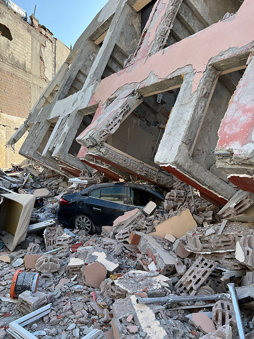 A building in Turkey slants on its side and rubble covers a car after the February 2023 earthquake