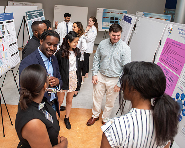 Students smile and talk in front of a row of research posters at PCOM Georgia's Research Day 2023 event