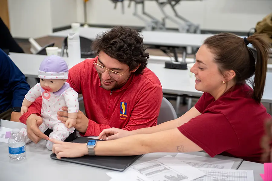 Two prospective physical therapy students smile as they work with an infant mannequin during a learning exercise