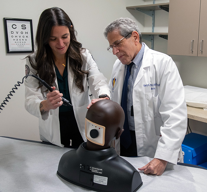 Dr. Freed instructs a medical student using an otoscope on a mannequin in an exam room