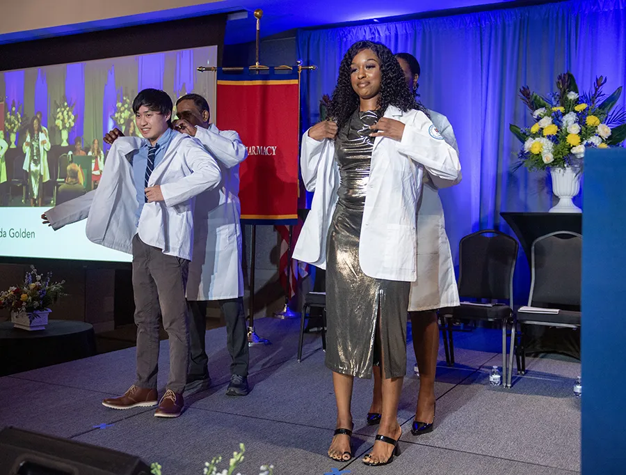 Two students smile as they don their white coats on a stage during the PCOM Georgia 2025 white coat ceremonies