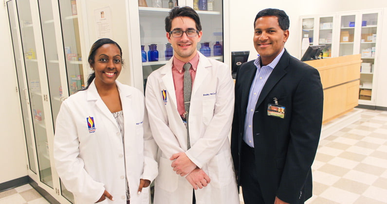 Megan Boudouin, PharmD, and Bradley McCoul, PharmD, wearing their white coats, stand beside program director Samuel John, PharmD, BCPS