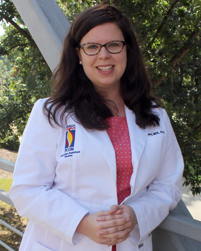 Carrie Smith Nold, MPA, PA-C, smiles as she stands outside on a walkway wearing her physician assistant white coat