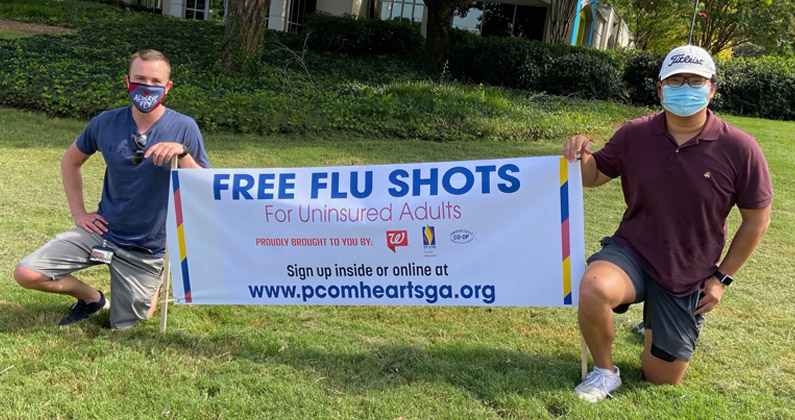 Two PCOM Georgia medical students wearing face masks kneel in front of a health clinic and hold a large banner promoting free flu shots for uninsured adults