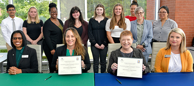Faculty, administrators and students from Georgia Gwinnett College and PCOM Georgia smile behind tables as part of the affiliation agreement signing event