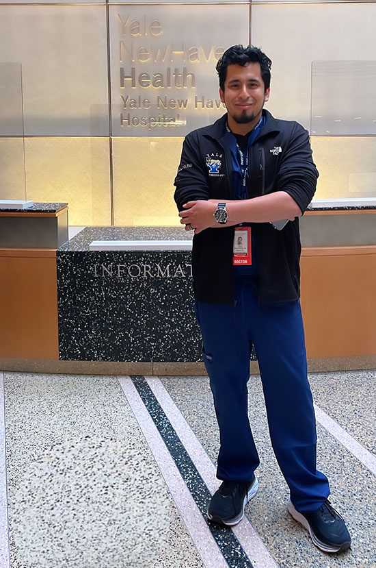PCOM graduate David R. Garcia Castro poses in front of a sign showing Yale New Haven Health
