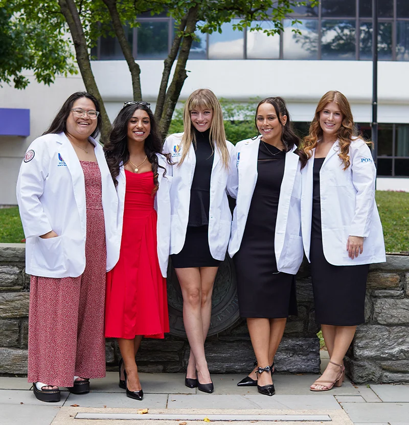 A group of female PCOM medical students smile on the Philadelphia campus grounds in their new student physician white coats