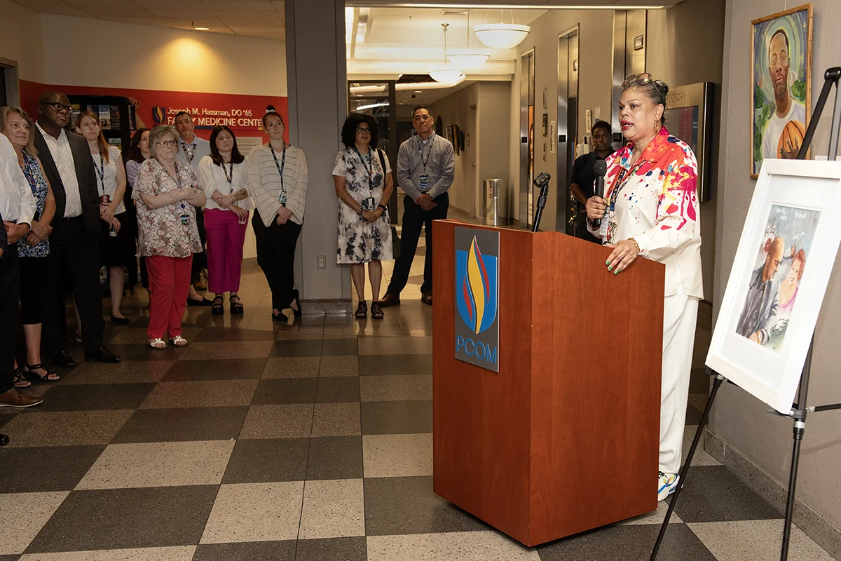 PCOM students and staff listen to a speaker at a podium opening the  Souls Shot Portrait Project art exhibit