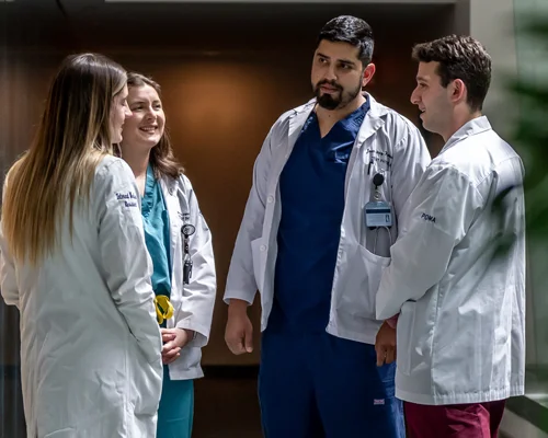 A group of medical students in white lab coats stand and talk in a hallway