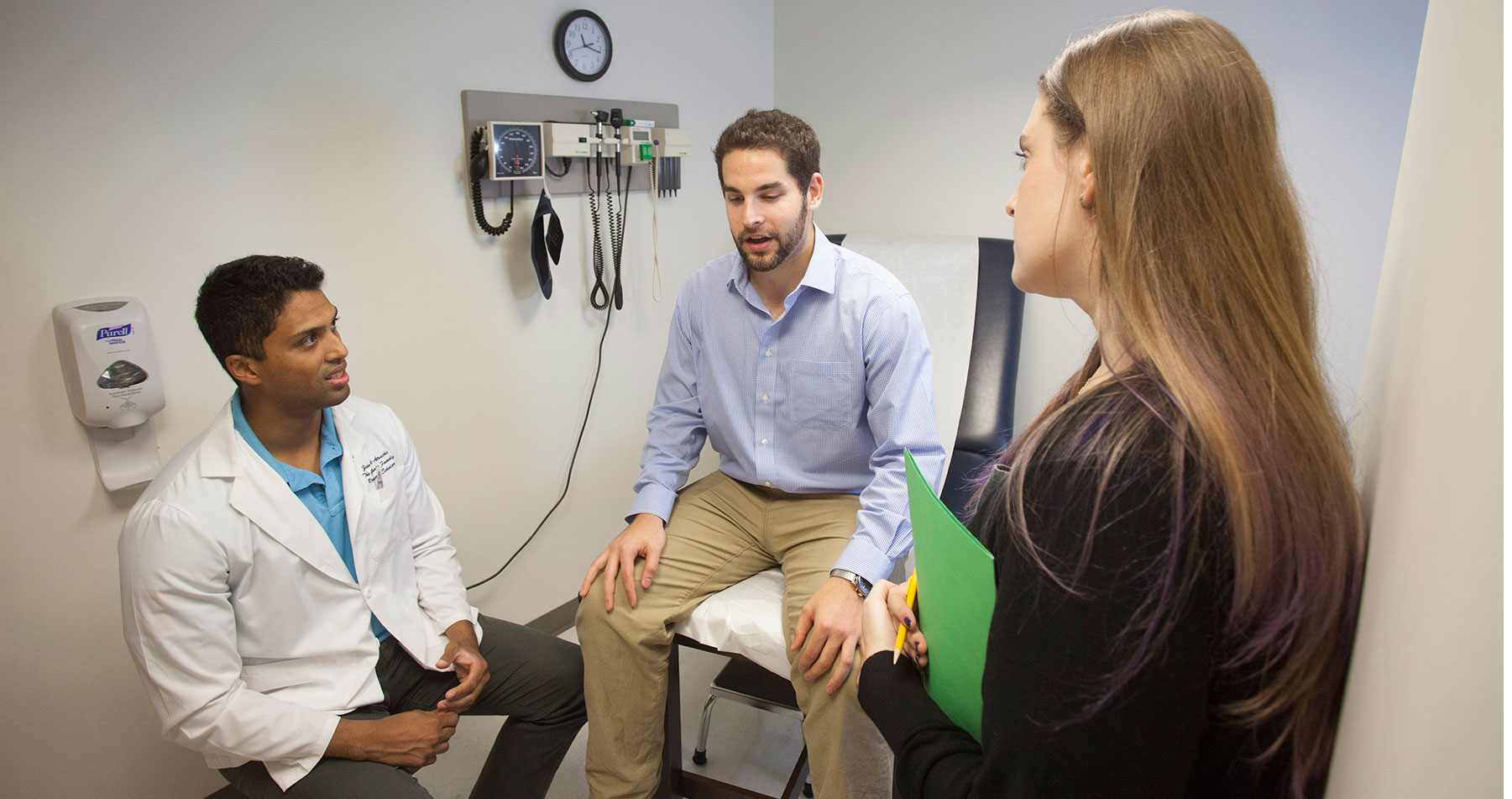 A PCOM medical student and psychology student speak with a patient actor in an exam room.