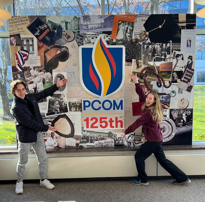 PCOM students pose in front of window-sized collage depicting moments from PCOM's history