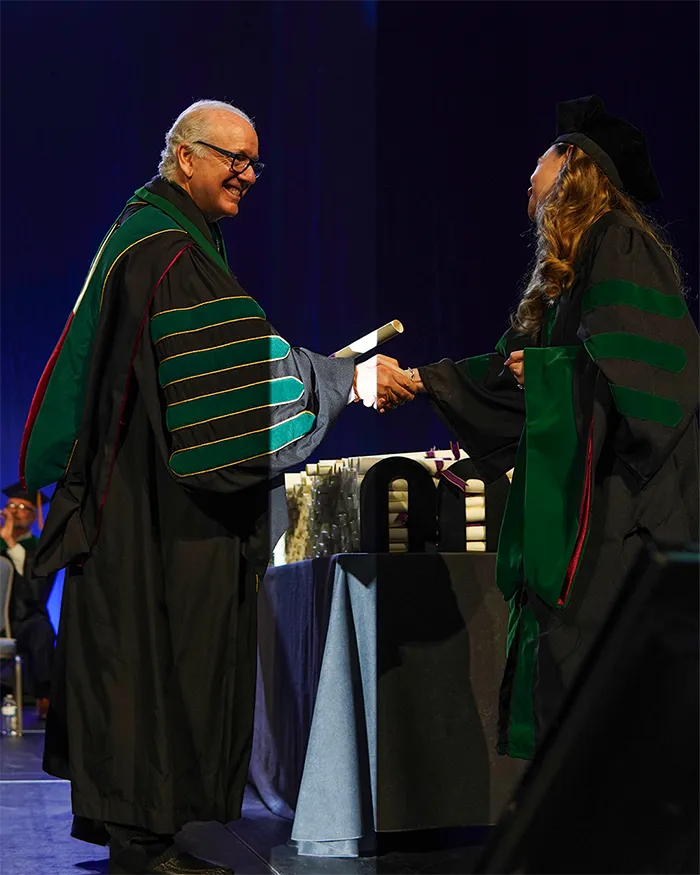 PCOM President Jay Feldstein, DO '81, shakes hands with a med school graduate as he hands them a diploma on stage during a 2025 commencement ceremony