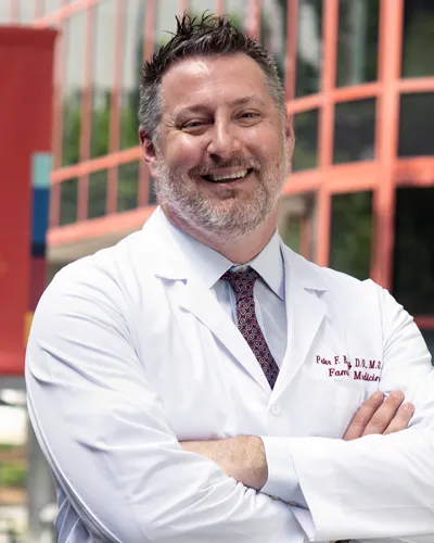 Professional headshot photograph of Peter Bidey, DO '08, MSEd, FACOFP in his physician white coat standing outside of PCOM's Philadelphia campus