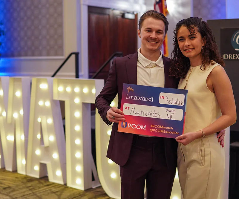 Young couple stands together in formal wear while one holds a "Match Day" sign