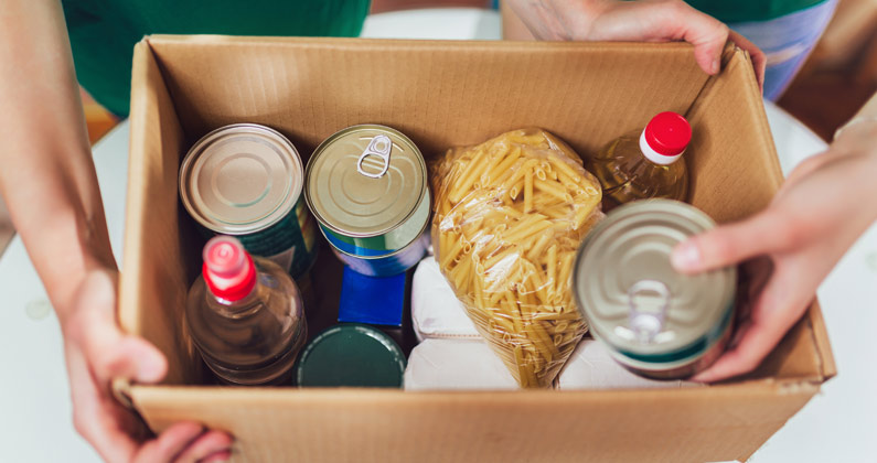 Hands holding a box full of food pantry items including fresh fruits and vegetables, canned goods and bagged pasta.