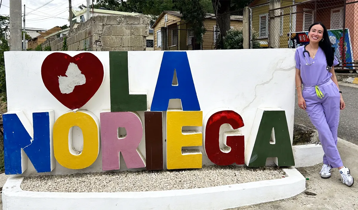 PCOM DO student Arlin Arias smiles next to colorful stone sign of her hometown Noriega in the Dominican Republic.